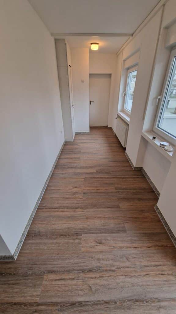 Ein schmaler, leerer Raum mit weißen, vom Malerfachbetrieb Stein gestrichenen Wänden, einem Holzboden, zwei großen Fenstern auf der rechten Seite, einer Deckenleuchte und einer geschlossenen Tür am anderen Ende. Durch die Fenster fällt natürliches Licht ein.